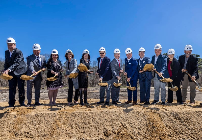A group of people participating in the AOE groundbreaking; everyone is smiling, holding a shovel with dirt and wearing white hardhats emblazoned with the AOE logo.